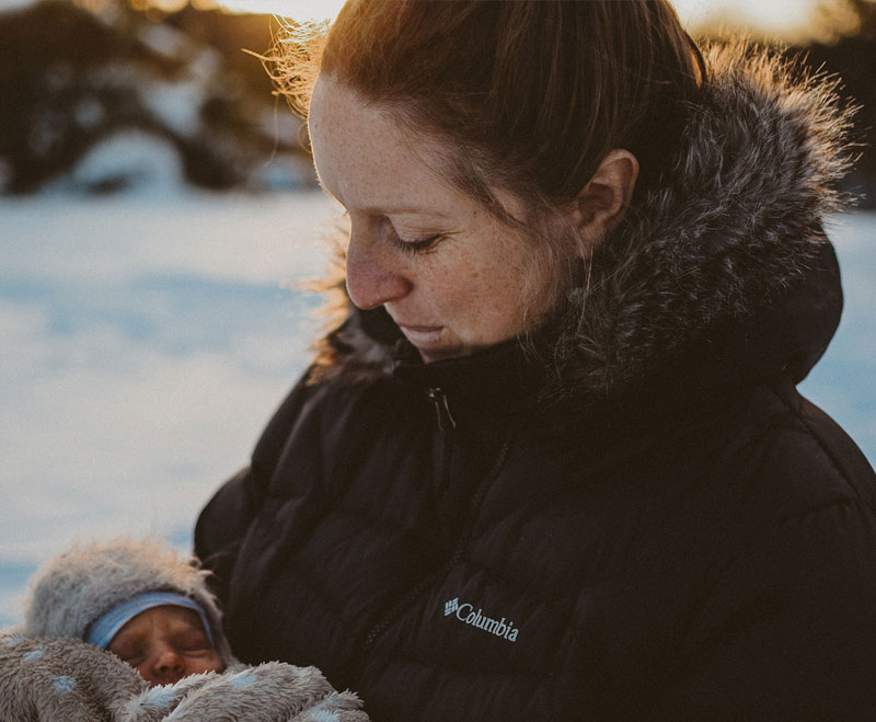 A new mother holds her baby bundled up outside with snow and ice in winter and the sun shining behind them A new mother holds her baby bundled up outside with snow and ice in winter and the sun shining behind them