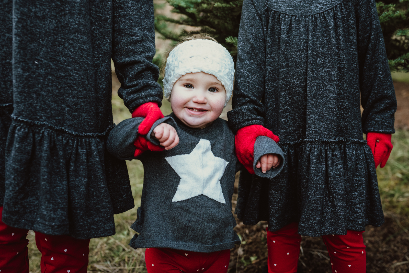 One year old girl stands outside with both her hands held by her two older sisters One year old girl stands outside with both her hands held by her two older sisters